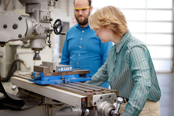 An NICC CNC student works at a lathe in the lab while a professor supervises.