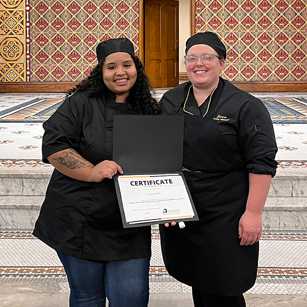 Alaina Miller holding her Culinary Foundations certificate, pictured next to an NICC instructor.
