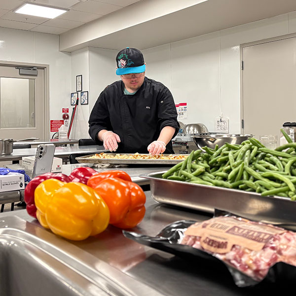 A chef chops ingredients in the background with peppers and beef on the table in front of him.