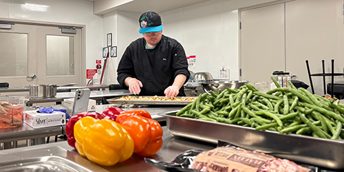 A chef chops ingredients in the background with peppers and beef on the table in front of him.
